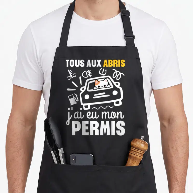 Tablier de cuisine noir avec illustration d'un chat au volant et texte tous aux abris j'ai eu mon permis en blanc et orange.