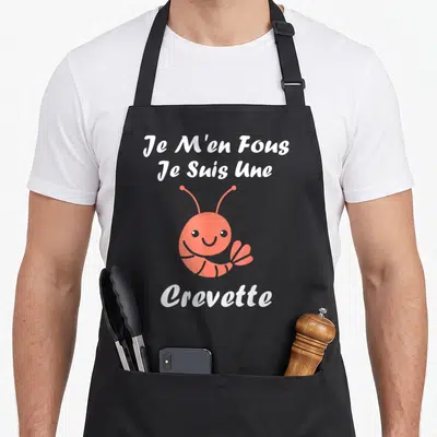 Tablier de cuisine noir avec une crevette rose souriante et le texte blanc Je M'en Fous Je Suis Une Crevette imprimé au centre.
