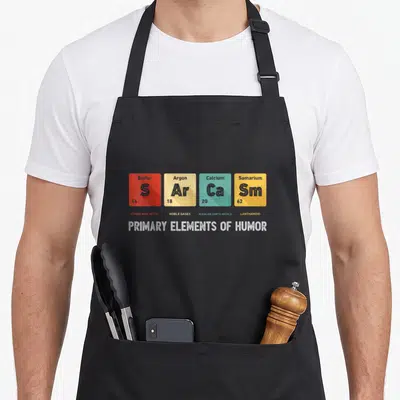 Tablier de cuisine noir avec quatre cases colorées imitant un tableau périodique chimique et le texte Primary Elements of Humor en bas.