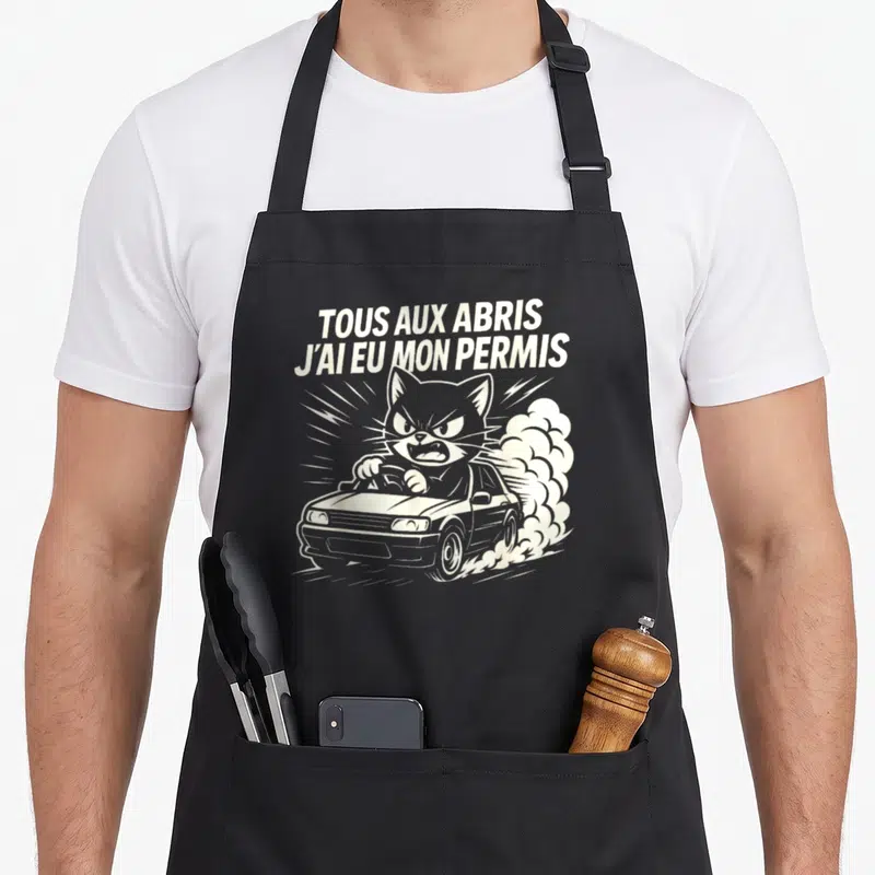 Tablier de cuisine noir avec illustration d'un chat agressif au volant et texte blanc Tous aux abris j'ai eu mon permis.