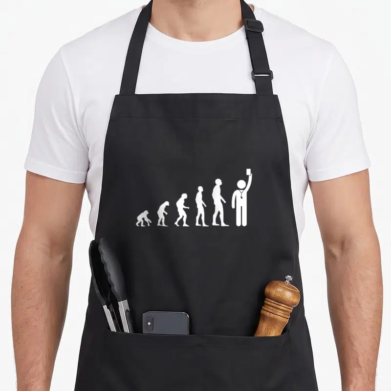 Tablier de cuisine noir avec illustration blanche représentant l'évolution humaine se terminant par un arbitre de football levant une carte.