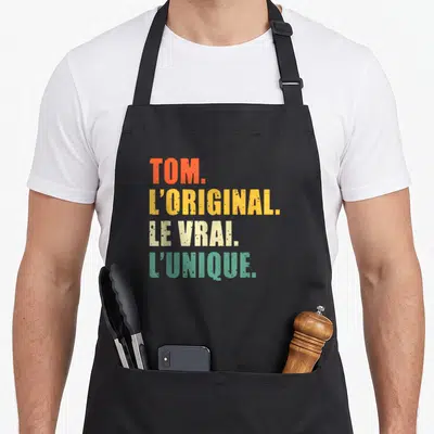 Tablier de cuisine noir avec les mots Tom l'original le vrai l'unique imprimés en couleurs vives.