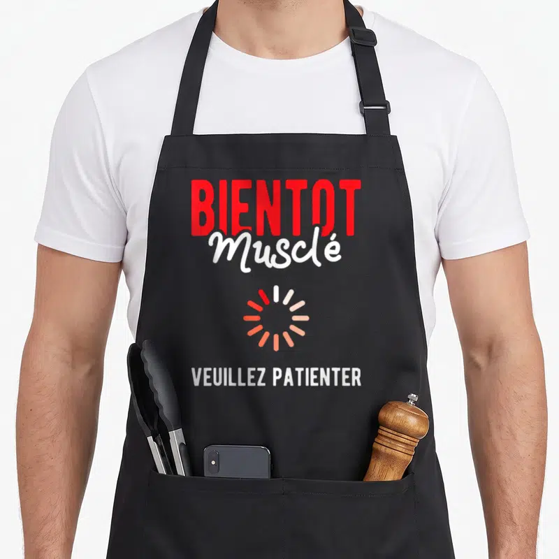 Tablier de cuisine noir avec l'inscription Bientôt Musclé Veuillez Patienter et un cercle de chargement coloré au centre.