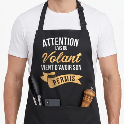 Tablier de cuisine noir avec inscription Attention l'as du volant vient d'avoir son permis en typographie blanche et orange sur bandeau.