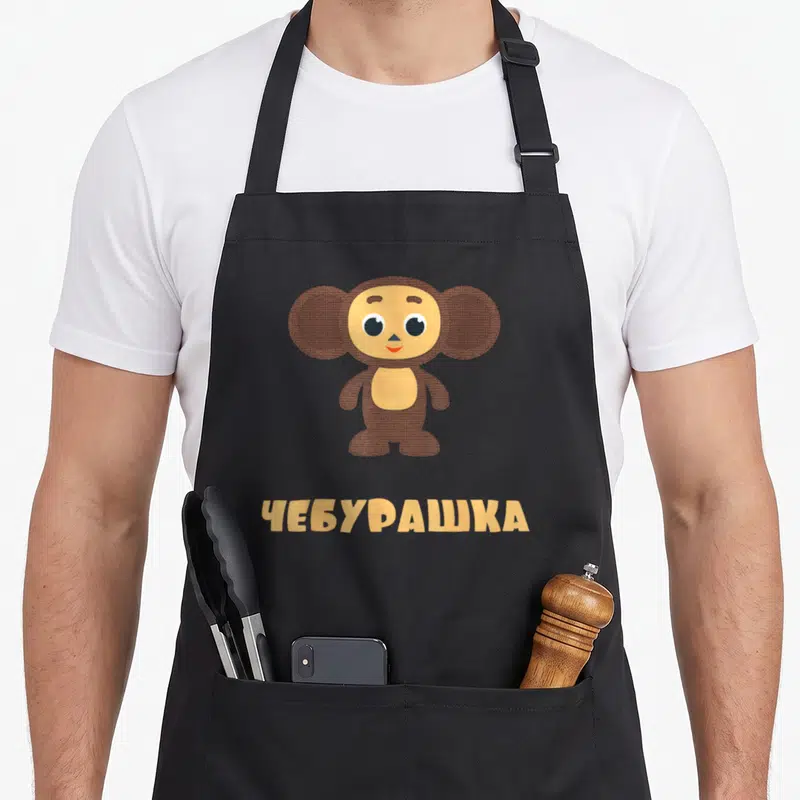 Tablier de cuisine noir avec un singe marron aux grands yeux et une inscription jaune en alphabet russe.