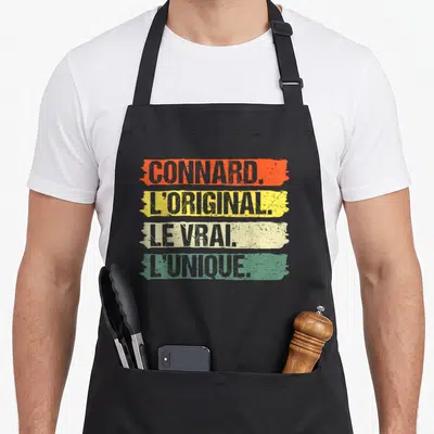 Tablier de cuisine noir avec les inscriptions Connard, L'Original, Le Vrai et L'Unique sur des fonds colorés délavés.