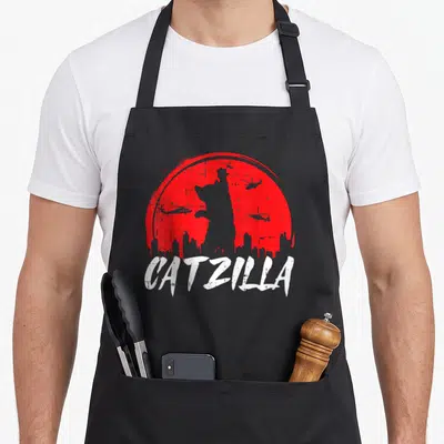 Tablier de cuisine noir avec silhouette de chat géant rouge et hélicoptères sur fond urbain.