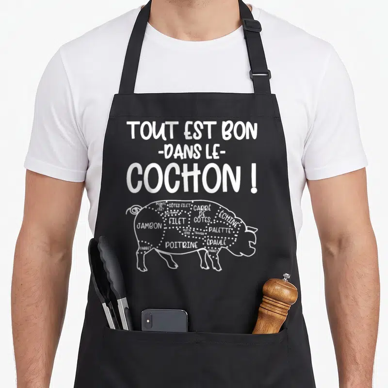 Tablier de cuisine noir avec texte tout est bon dans le cochon et illustration de découpe porcine en blanc.
