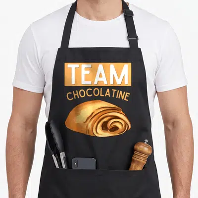 Tablier de cuisine noir avec illustration d'un pain au chocolat et texte team chocolatine en orange et blanc.