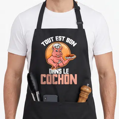 Tablier de cuisine noir avec illustration d'un porc musclé en toque tenant une assiette de saucisses et une fourche.