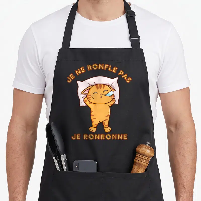 Tablier de cuisine noir avec illustration d'un chat tigré doré dormant sur un oreiller blanc, texte orange en haut et en bas.