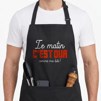 Tablier de cuisine noir avec l'inscription Le matin c'est dur comme ma bite en lettres rouges et blanches.