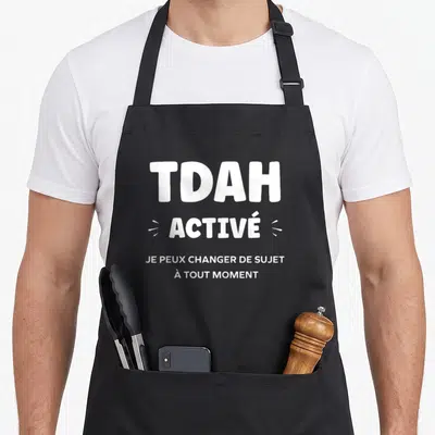 Tablier de cuisine noir avec l'inscription blanche TDAH ACTIVÉ et une phrase explicative humoristique centrée sur le motif.