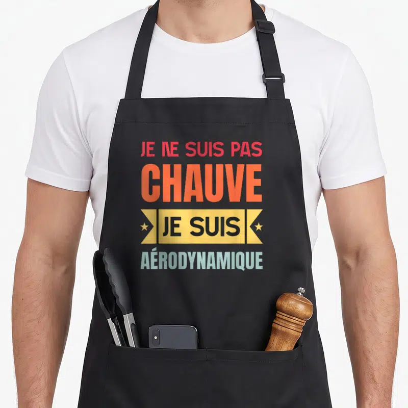 Tablier de cuisine noir avec l'inscription Je ne suis pas chauve je suis aérodynamique en lettres orange et jaune.