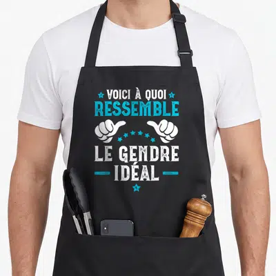 Tablier de cuisine noir avec inscription Voici à quoi ressemble le gendre idéal et deux mains blanches pointant vers le texte central.