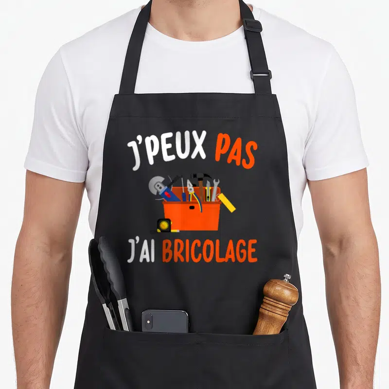 Tablier de cuisine noir avec une boîte à outils orange et l'inscription J'peux pas j'ai bricolage en lettres blanches et oranges.