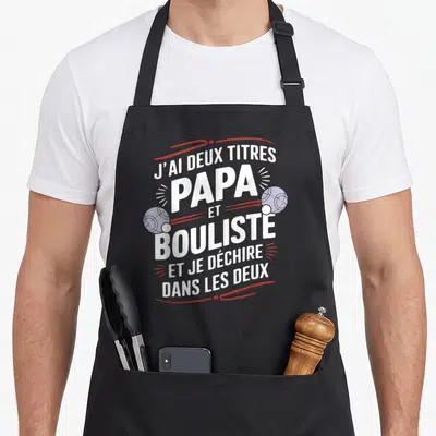 Tablier de cuisine noir avec inscription blanche Papa et Bouliste et deux boules de pétanque grises sur fond gris.