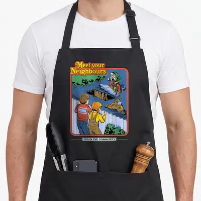 Tablier de cuisine noir avec illustration rétro Meet your Neighbours montrant un fossé derrière une clôture blanche.
