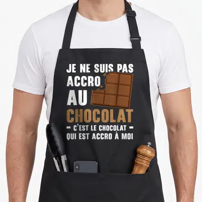Tablier de cuisine noir avec texte blanc et marron sur fond gris et illustration d'une barre de chocolat mordue.