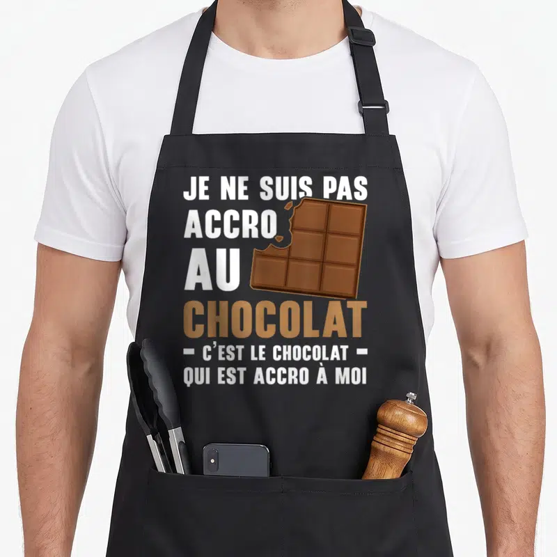 Tablier de cuisine noir avec texte blanc et marron sur fond gris et illustration d'une barre de chocolat mordue.