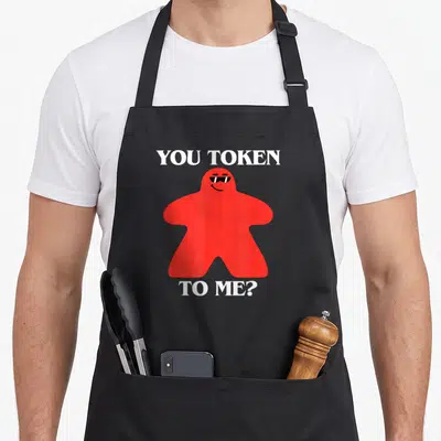 Tablier de cuisine noir avec un meeple rouge portant des lunettes noires et du texte blanc humoristique.