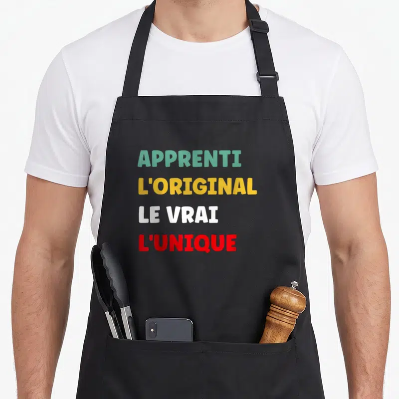 Tablier de cuisine noir avec inscription Apprenti L'Original Le Vrai L'Unique en lettres vertes jaunes blanches et rouges.