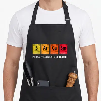 Tablier de cuisine noir avec quatre cases colorées imitant une table de périodicité chimique sur fond gris uni.