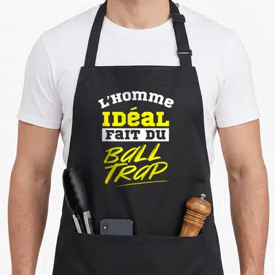 Tablier de cuisine noir avec le texte L'homme idéal fait du ball trap imprimé en jaune et blanc.