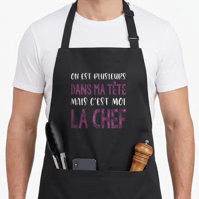 Tablier de cuisine noir avec le texte On est plusieurs dans ma tête mais c'est moi la chef en rose et blanc.