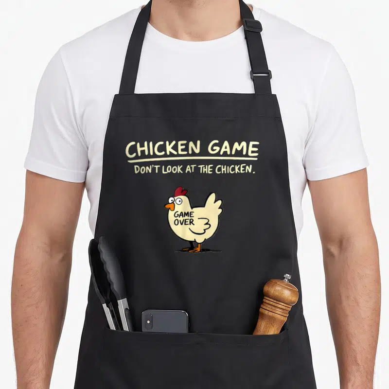 Tablier de cuisine noir avec illustration d'un poulet blanc aux yeux ronds et texte chicken game en lettres capitales jaunes.
