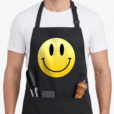 Tablier de cuisine noir avec un grand smiley jaune aux yeux noirs et sourire large.