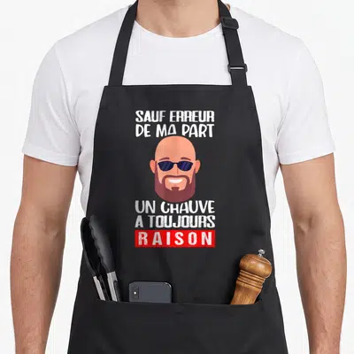 Tablier de cuisine noir avec illustration d'un homme chauve barbu portant des lunettes de soleil et du texte humoristique en blanc et rouge.