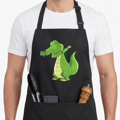 Tablier de cuisine noir avec un crocodile vert cartoon effectuant une pose dab amusante sur le devant.