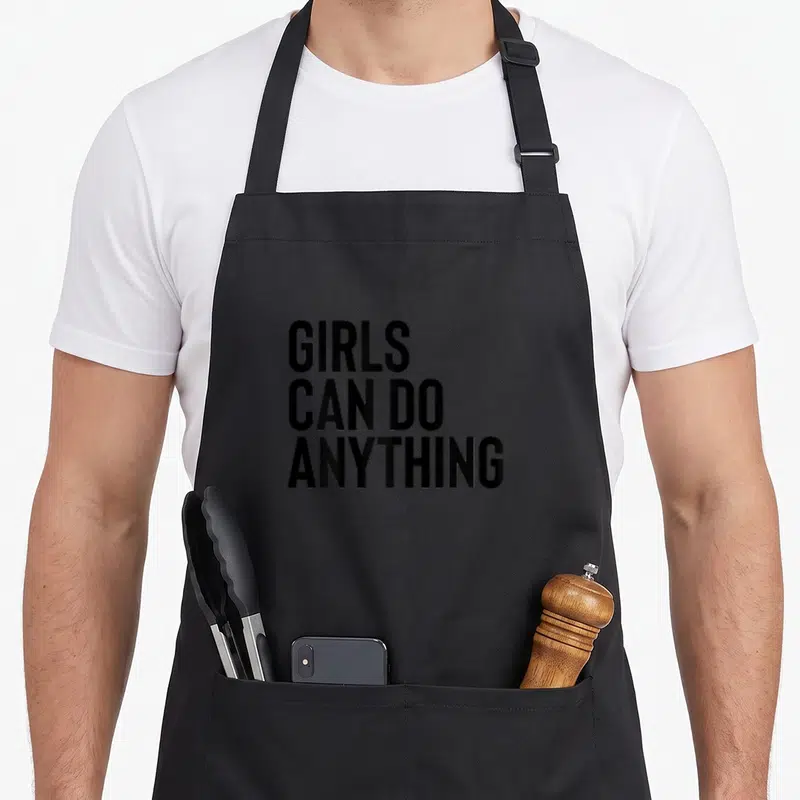 Tablier de cuisine noir avec l'inscriptionGirls Can Do Anything en lettres capitales noires au centre.