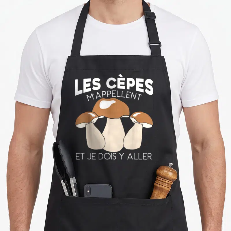 Tablier de cuisine noir avec illustration de trois cèpes et texte blanc humoristique sur fond uni.