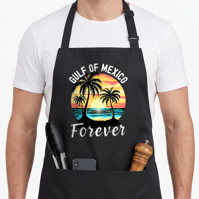 Tablier de cuisine noir avec illustration circulaire de palmiers et soleil couchant sur l'eau, texte Gulf of Mexico Forever en blanc.