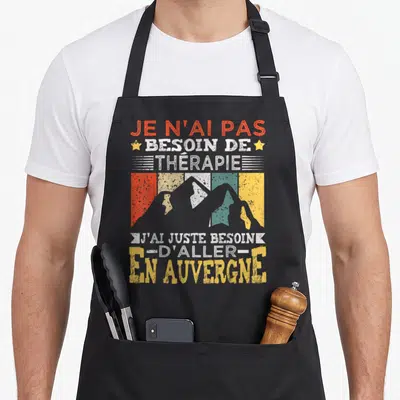 Tablier de cuisine noir avec inscription humoristique sur l'Auvergne et silhouettes de montagnes colorées style rétro.