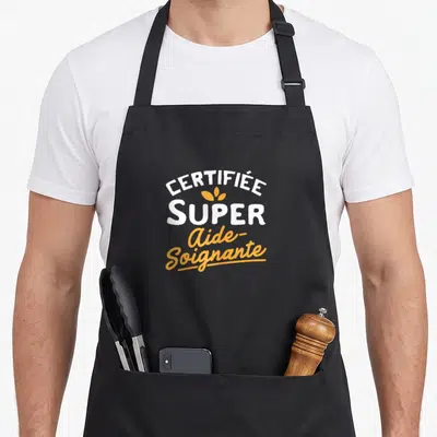 Tablier de cuisine noir avec linscription certifiée super aide-soignante en blanc et orange et une petite feuille stylisée au centre.