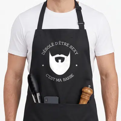 Tablier de cuisine noir avec une illustration blanche représentant une barbe stylisée et une moustache entre deux étoiles.