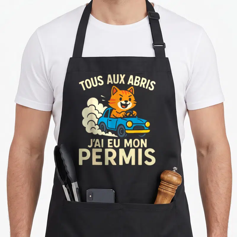 Tablier de cuisine noir avec un chat orange au volant d'une voiture bleue et le texte Tous aux abris j'ai eu mon permis.