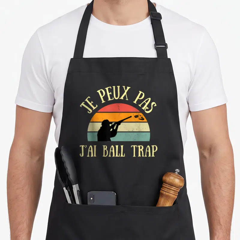 Tablier de cuisine noir avec silhouette tireur et texte Je peux pas j'ai ball trap sur fond de soleil coloré.
