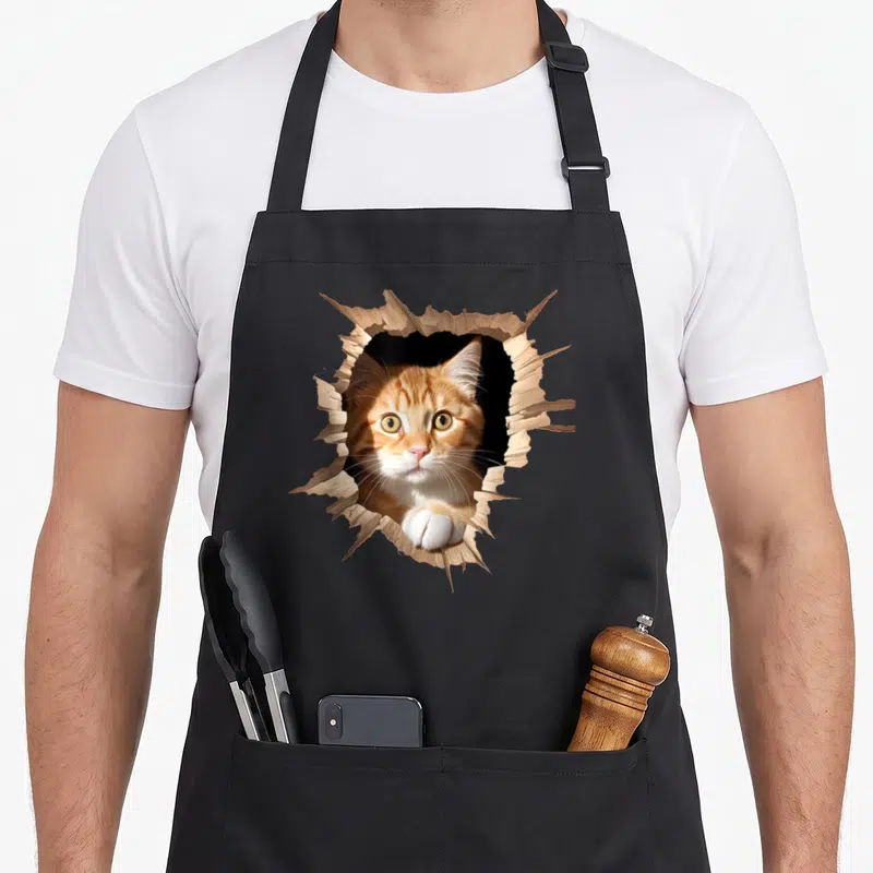 Tablier de cuisine noir avec un chat orange percant une surface en bois dans un effet trompe-l'œil 3D.