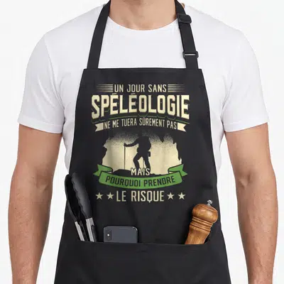 Tablier de cuisine noir avec silhouette spéléologue et texte humoristique sur fond beige et vert.