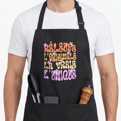 Tablier de cuisine noir avec inscription Râleuse L'Originale La Vraie L'Unique en typographie vintage colorée et petites fleurs blanches.