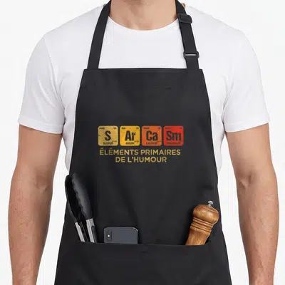 Tablier de cuisine noir avec motif tableau périodique coloré et texte éléments primaires de l'humour en jaune.
