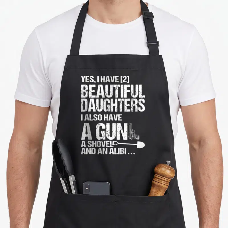 Tablier de cuisine noir avec inscription blanche « Yes I Have 2 Magnifiques Filles » et dessin d'une arme et d'une pelle.