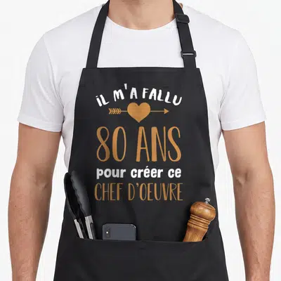 Tablier de cuisine noir avec inscription Il m'a fallu 80 ans pour créer ce chef d'oeuvre et un coeur orange traversé d'une flèche.