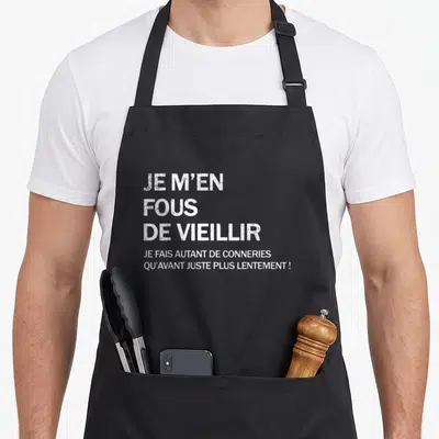 Tablier de cuisine noir avec texte blanc « Je m'en fous de vieillir » et une phrase humoristique en dessous.
