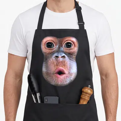Tablier de cuisine noir avec l'illustration d'un visage de singe soufflant le ventre en gros plan.