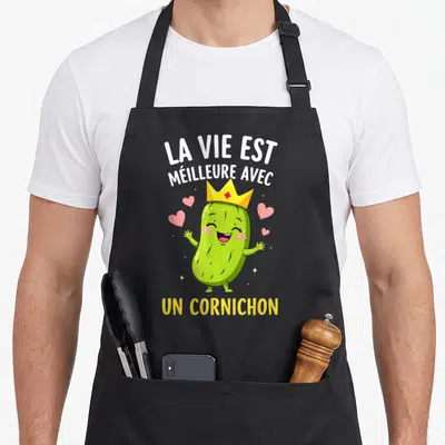 Tablier de cuisine noir avec un cornichon vert souriant portant une couronne jaune et des cœurs roses flottants.